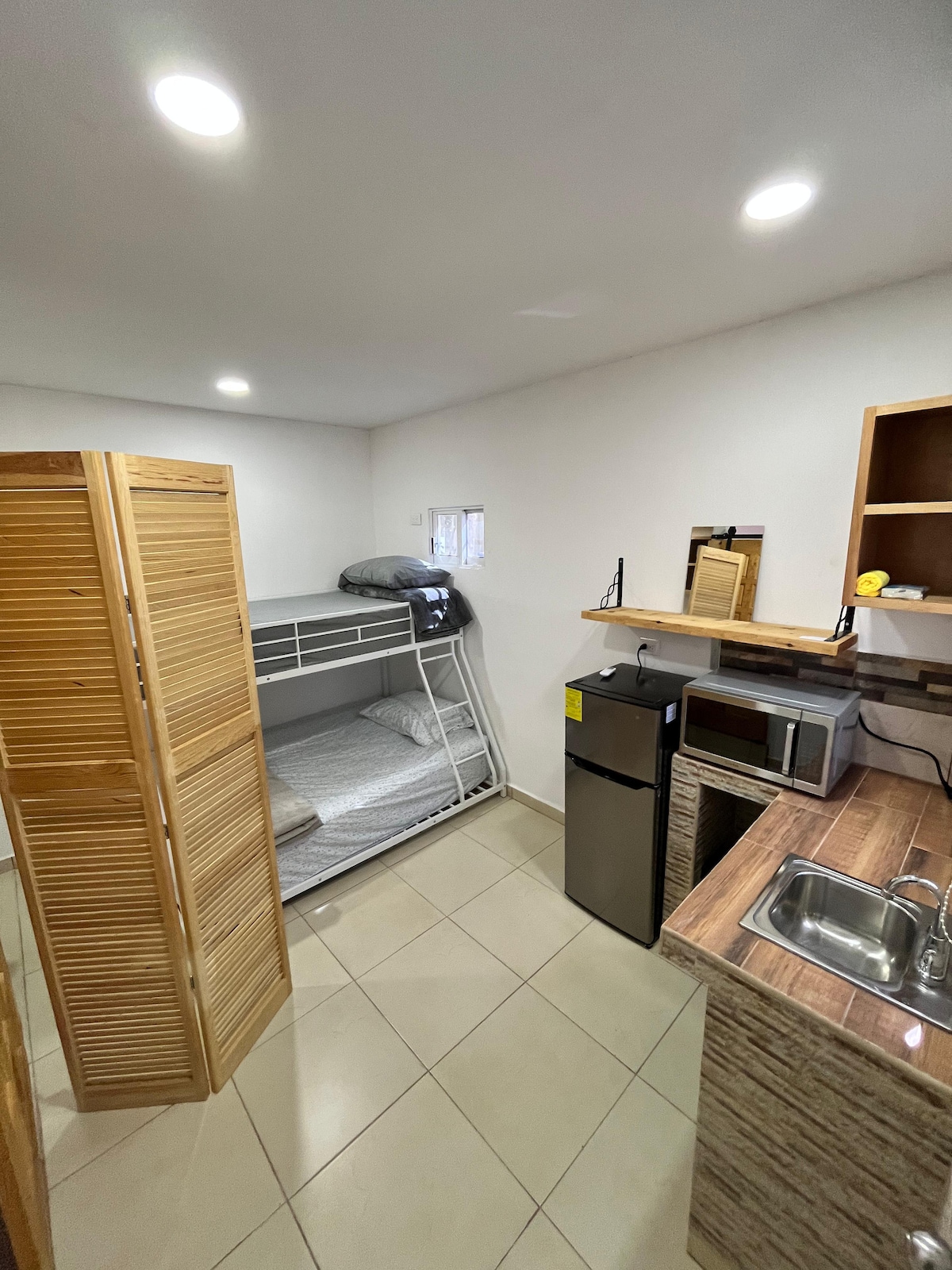 A functional space is highlighted by a wooden screen dividing the sleeping area from the kitchenette. A bunk bed is present, with clean linens and two positioned mattresses. The kitchenette includes a sink, microwave, and a compact refrigerator, with tile flooring and bright overhead lighting.