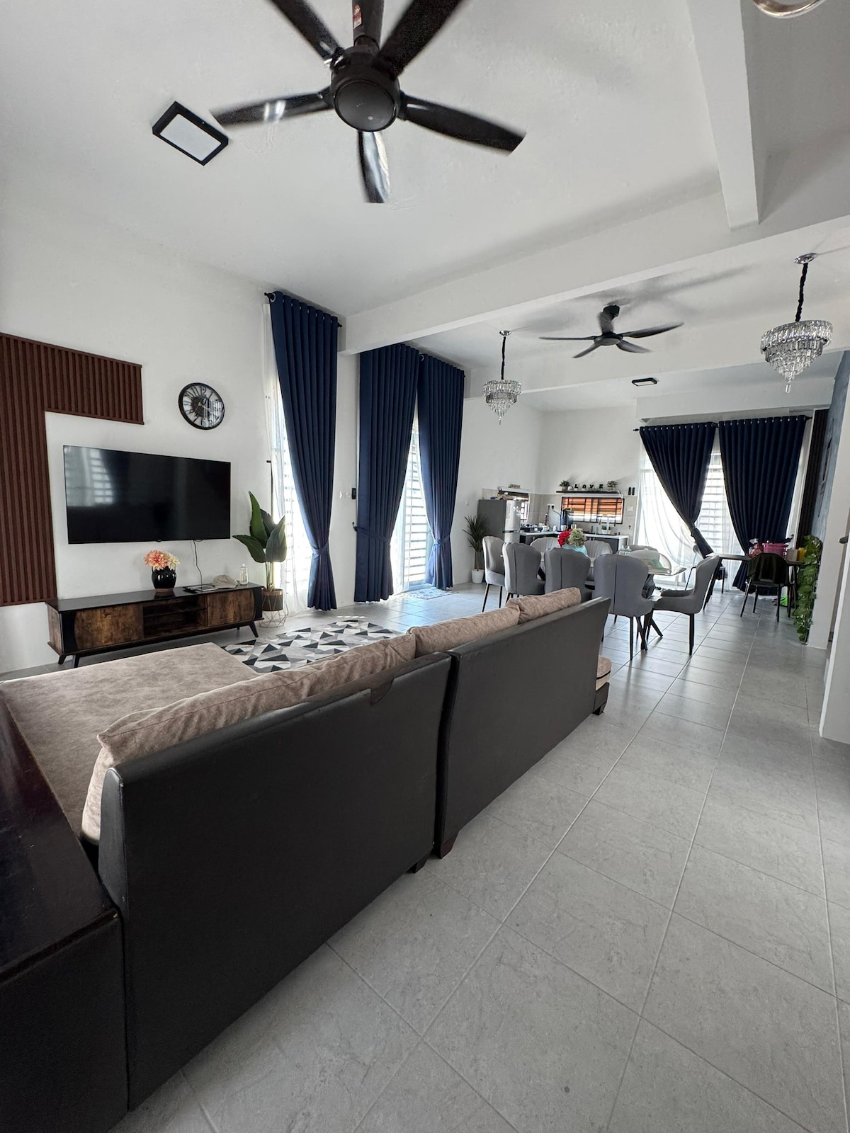 A spacious living area features a modern sectional sofa facing a flat-screen television mounted on the wall. Floor-to-ceiling windows are framed by elegant navy curtains, allowing natural light to enter. A dining area with stylish chairs and a chandelier is visible in the background.