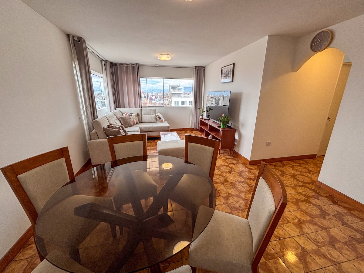 An open-concept living area is presented, featuring a glass dining table surrounded by six padded chairs. A comfortable sofa is positioned near large windows with light drapes, providing ample natural light, and a television stand is seen against the wall.