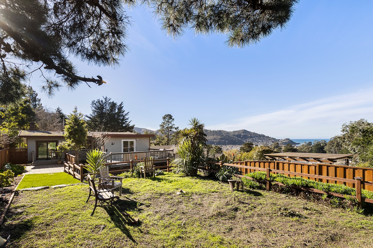 The expansive backyard is surrounded by lush vegetation and features manicured grass. A wooden deck is visible with seating options. In the distance, rolling hills and the coastline create a tranquil backdrop under a clear blue sky.