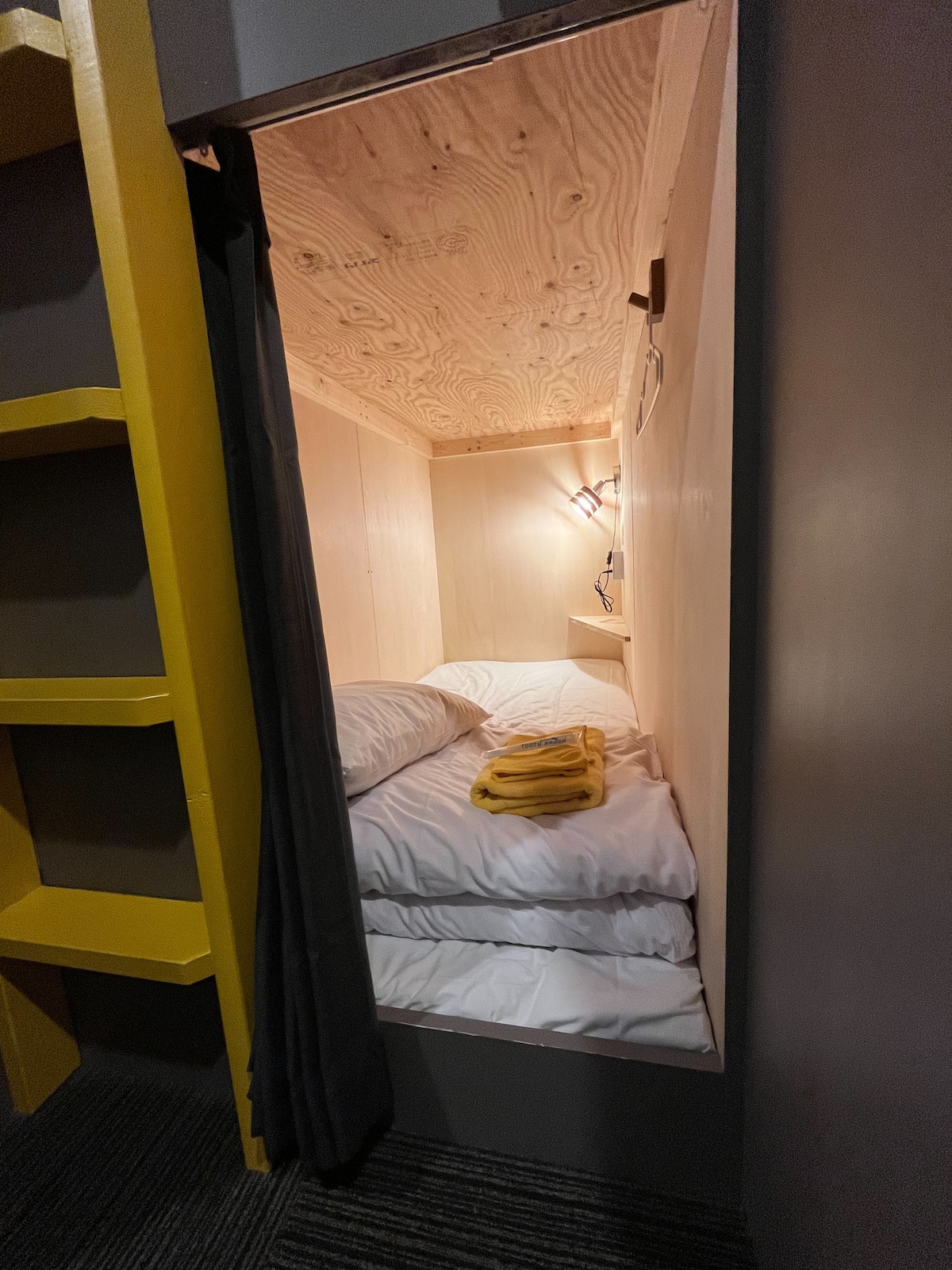 A capsule sleeping area is illuminated by soft lighting, featuring two neatly arranged mattresses with white bedding. A set of yellow towels is placed on top. The space is enclosed by wooden walls, providing a cozy and private atmosphere.