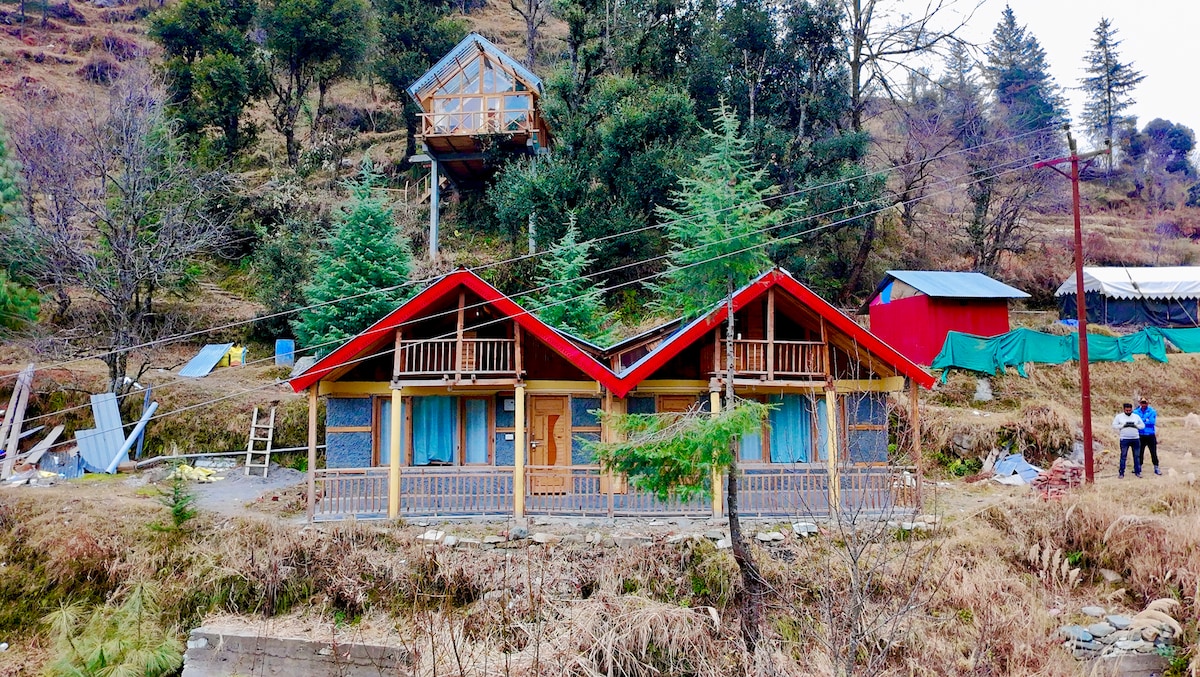 Two identical duplex cottages with slanted red roofs are nestled among the trees. A wooden deck with railings is visible beneath large windows. Nearby, a wooden structure is perched on stilts, and greenery surrounds the area, enhancing the natural setting.