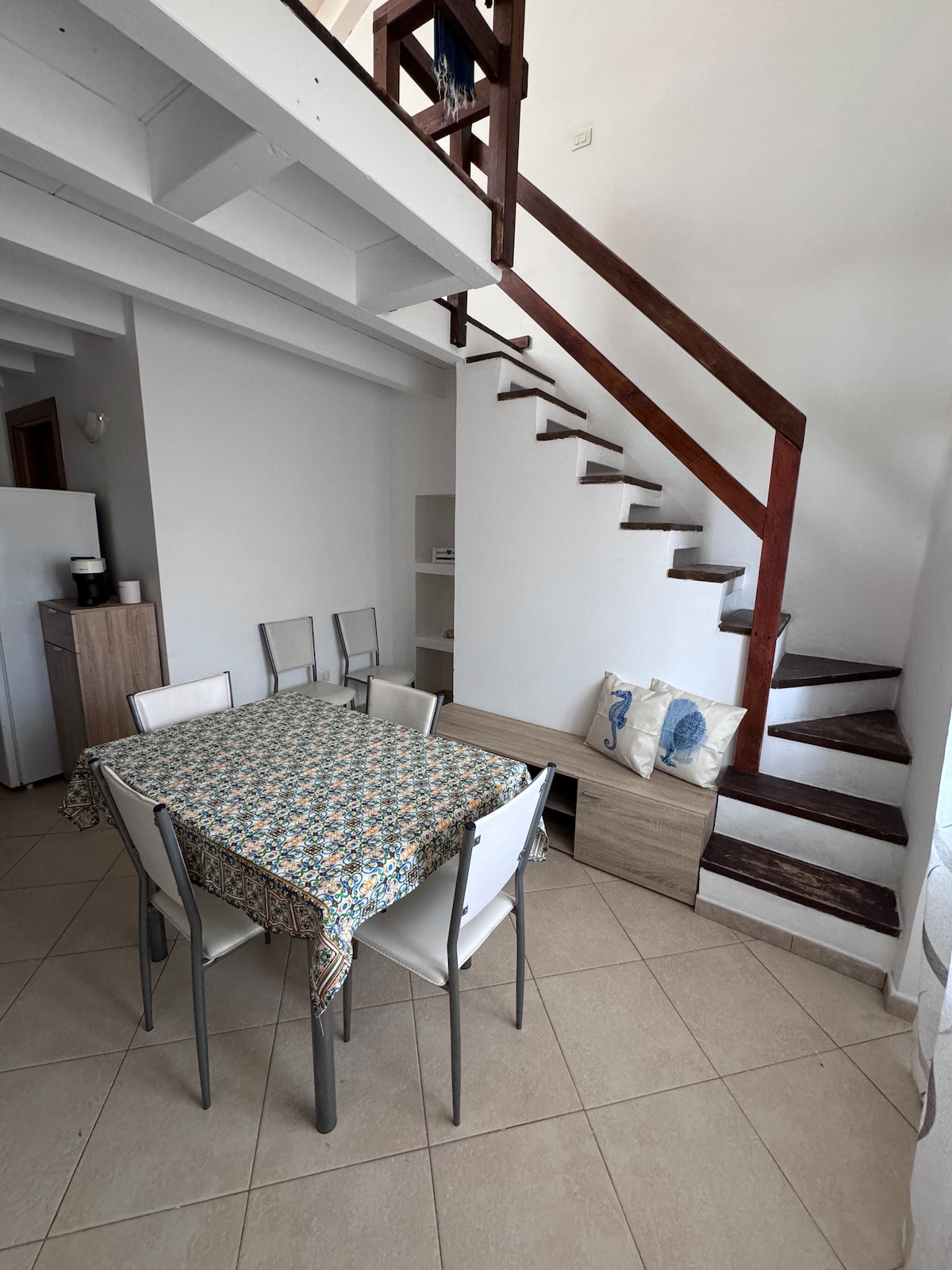 A dining area features a patterned tablecloth covering a rectangular table surrounded by six chairs. A staircase with wooden steps ascends alongside a light-colored wall, leading to a lofted space. Natural light fills the room, highlighting the neutral tones and clean lines.