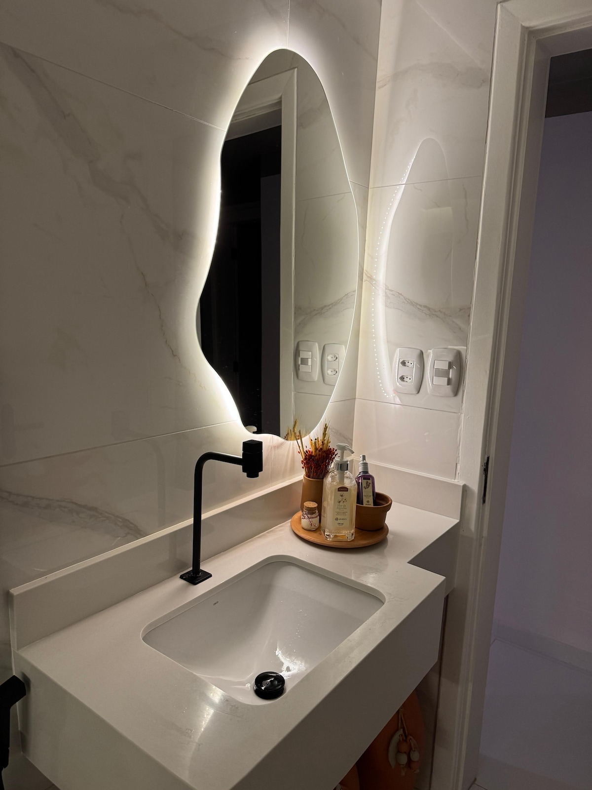 A modern bathroom sink area features a minimalist design with a smooth countertop and an integrated mirror illuminated by soft lighting. A wall-mounted faucet complements the elegant aesthetic, while small toiletries are neatly arranged beside a decorative vase, enhancing the functional yet stylish space.