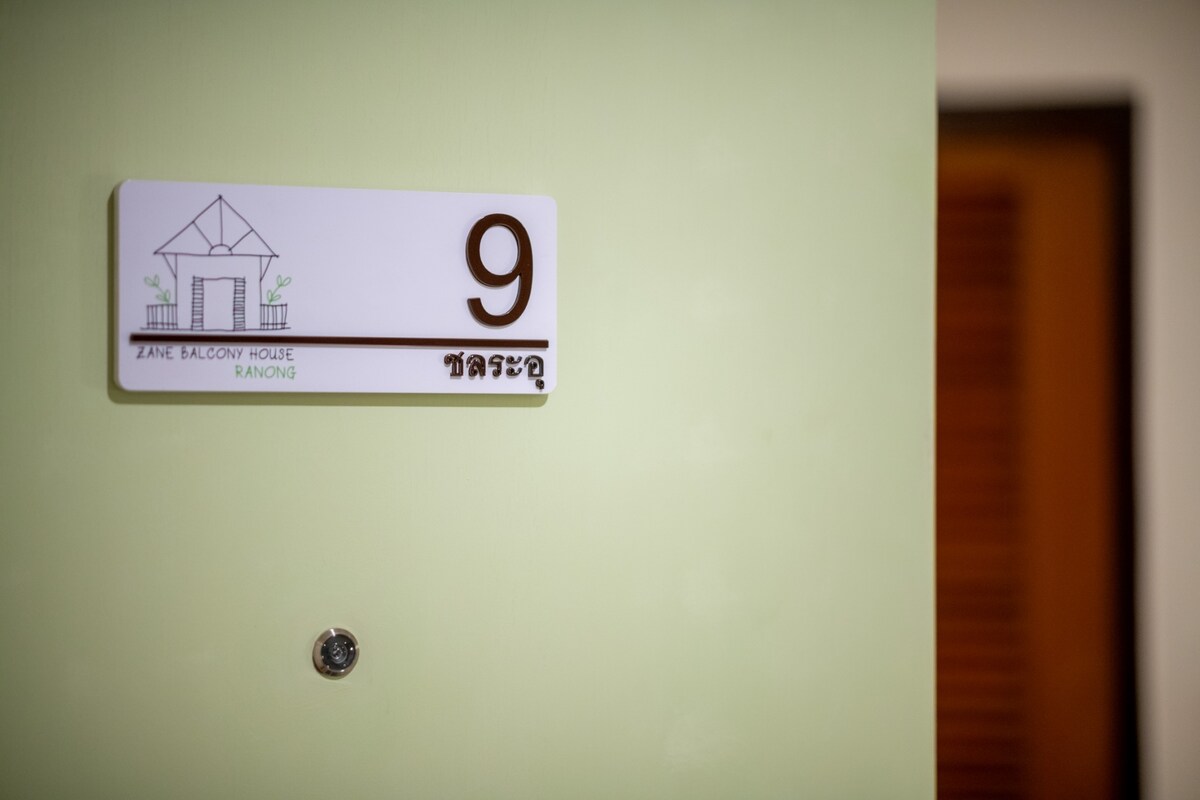 A room number sign is prominently displayed on a green wall, indicating room 9. The design is simple and modern, featuring a small house illustration above the text. A door is partially visible in the background.