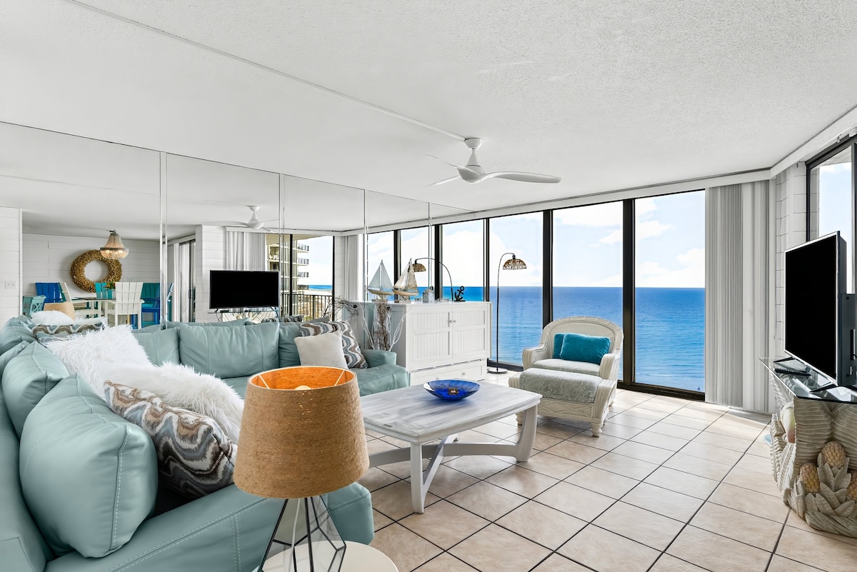 A spacious living area features a light blue sectional sofa complemented by a coffee table resting on tiled flooring. Large windows provide unobstructed ocean views, while a ceiling fan enhances airflow. Wall mirrors reflect the sunlight, adding brightness to the room.