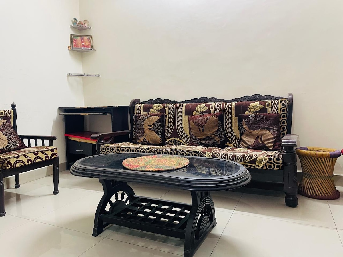 A seating area is shown, featuring a dark wooden sofa adorned with intricate patterns. A round coffee table sits in the center, accompanied by a small chair in a corner. Light-colored tiles enhance the room's brightness, while a floating shelf displays decorative items.