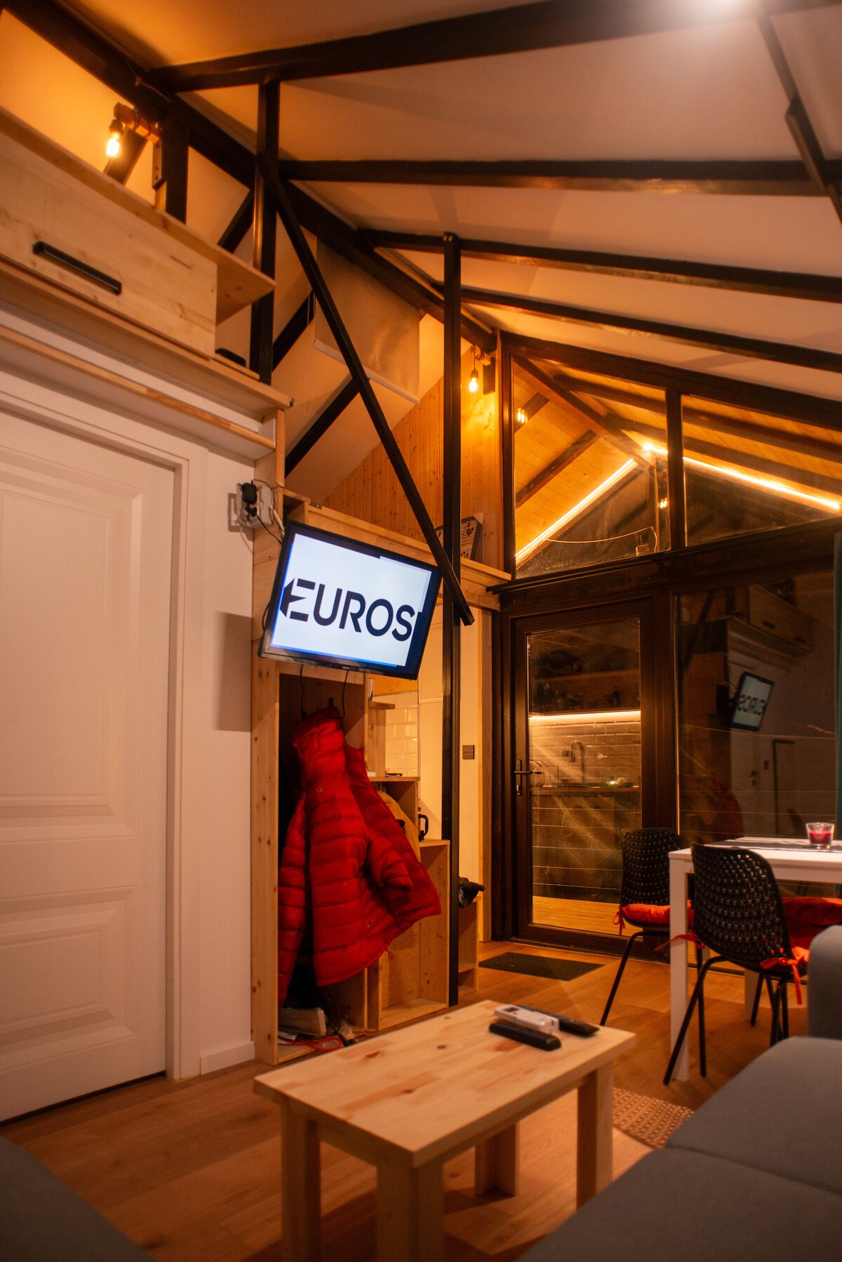 A modern interior space is highlighted by warm wood accents and industrial features. A flat-screen television is mounted on the wall near a cozy red jacket. Large windows provide ample natural light, while a small wooden table sits at the center with black dining chairs surrounding it.
