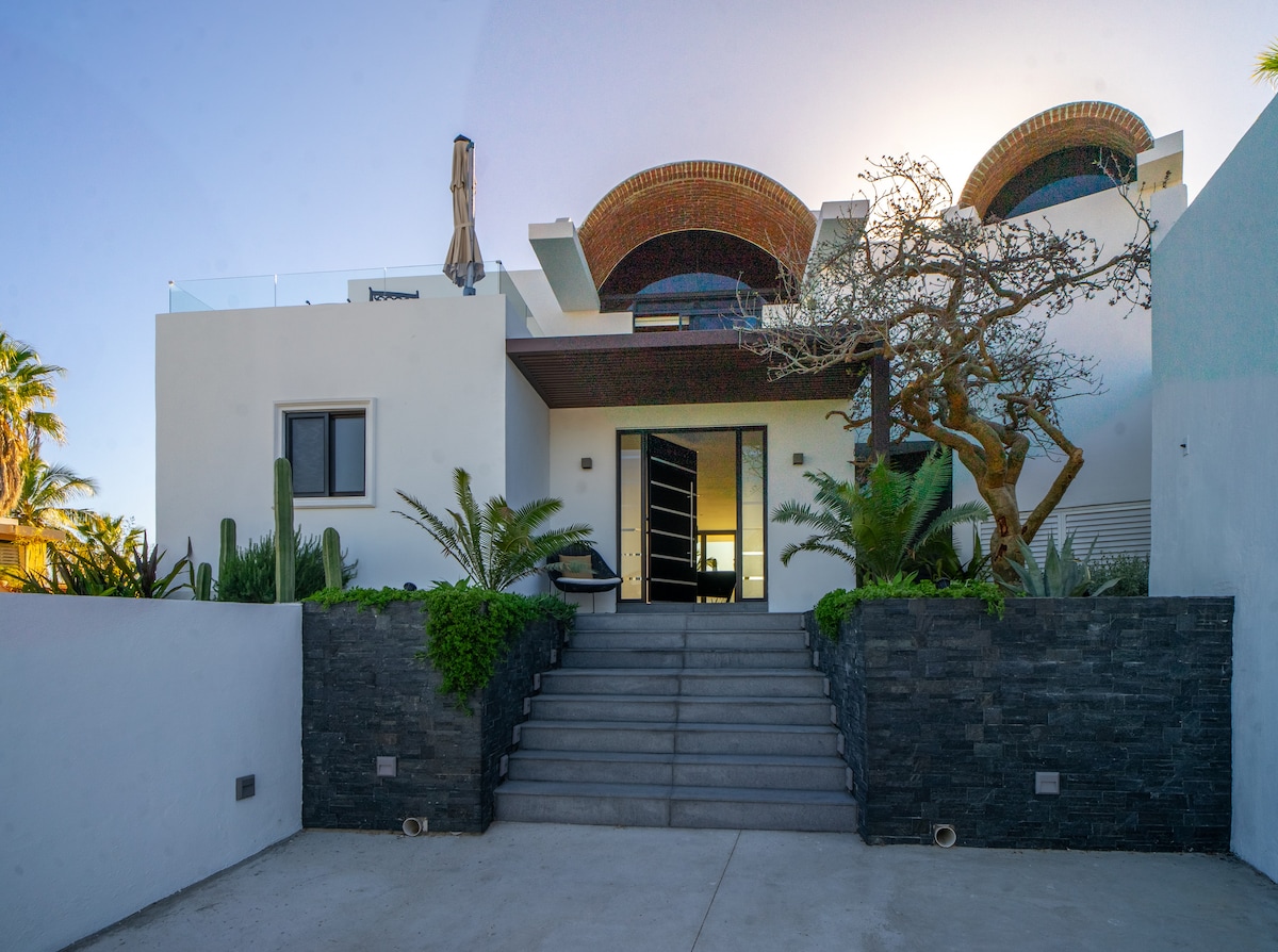 The villa entrance is framed by a series of steps leading up to a large glass door. Lush greenery, including palm and decorative plants, surrounds the pathway, while a striking tree adds visual interest to the space. Unique architectural elements can be seen in the roof design.