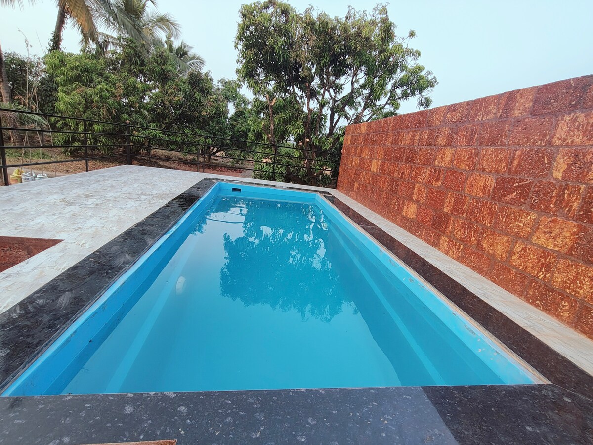 A clean swimming pool is set against a textured stone wall. Gazebo-style seating is visible nearby, surrounded by lush greenery and trees. The tranquil water reflects the open sky, creating a serene atmosphere.
