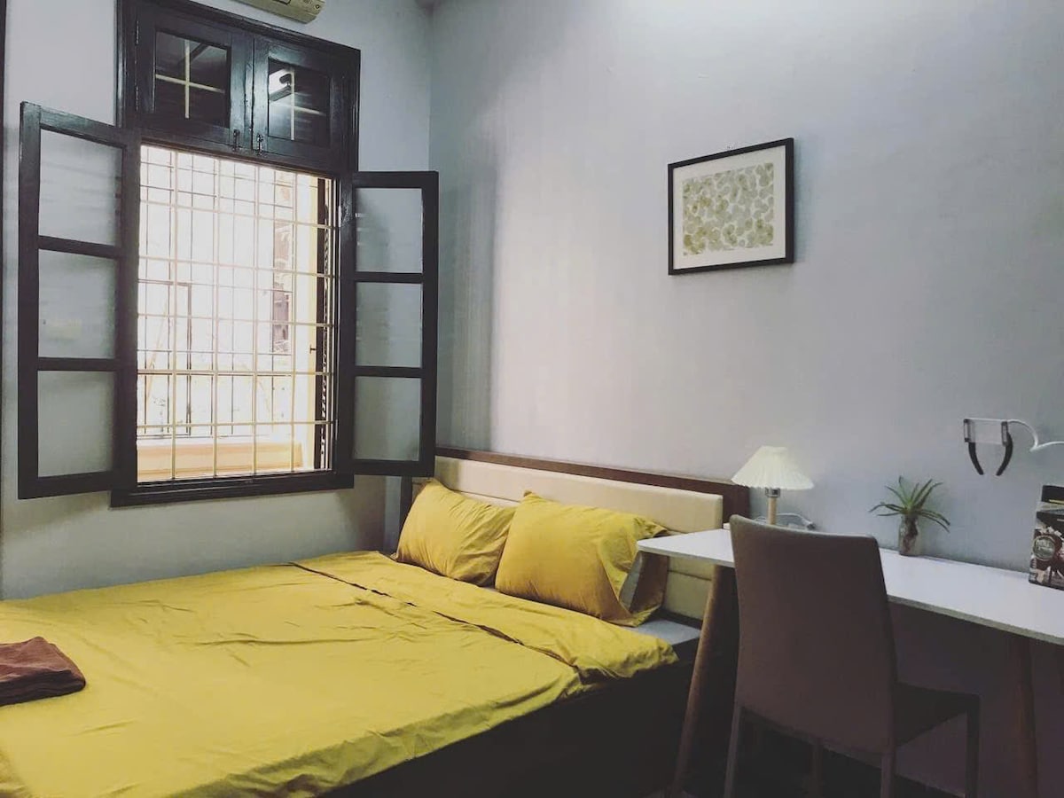 A sleeping area features a spacious bed with yellow bedding, complemented by a light-colored headboard. A small desk is positioned against the wall, equipped with a lamp and a decorative plant. A window with black shutters allows natural light to enter the room.