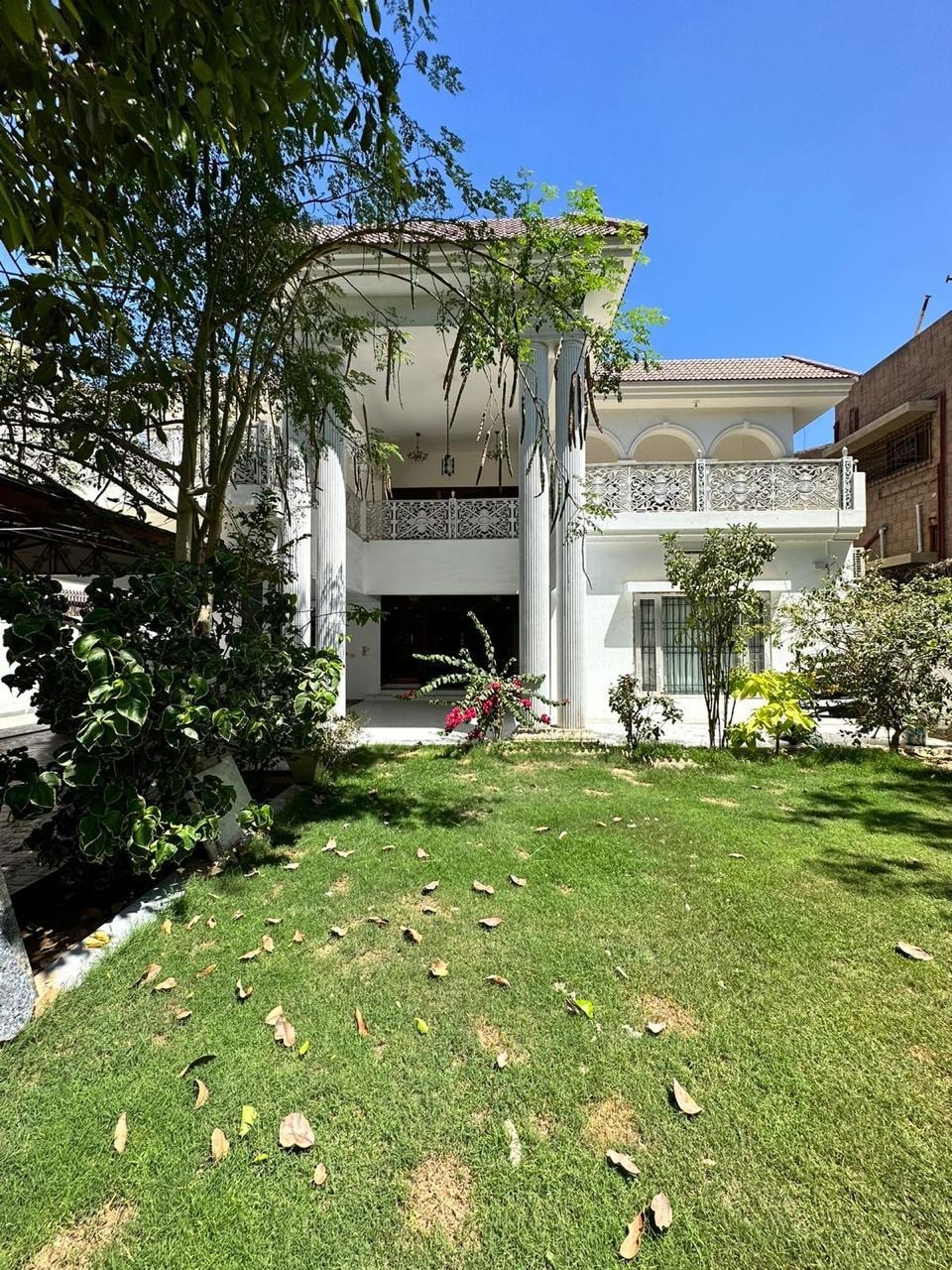The exterior of a two-story house is shown, featuring tall columns and arched balconies. Lush green grass surrounds the base, with scattered leaves on the ground. Colorful plants add natural beauty, while clear blue skies provide a bright backdrop.