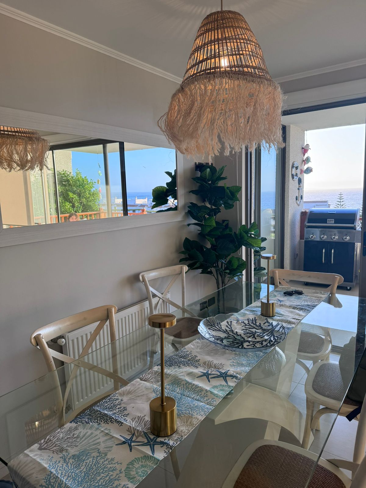 A dining area is presented with a clear glass table surrounded by white chairs. A decorative table runner features a blue and white pattern. A pendant light made of natural fibers hangs above. Large windows offer views of the ocean and provide ample natural light.