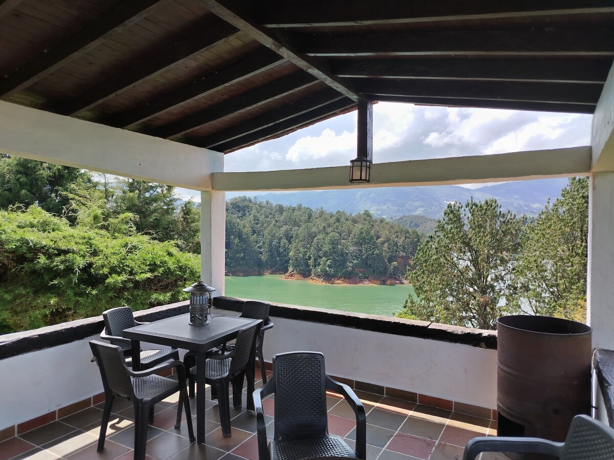 An outdoor terrace is presented with a dark wooden ceiling. A black table surrounded by four chairs overlooks lush greenery and a turquoise reservoir. Natural light filters through the open space, creating a serene spot for relaxation and enjoying the surrounding views.