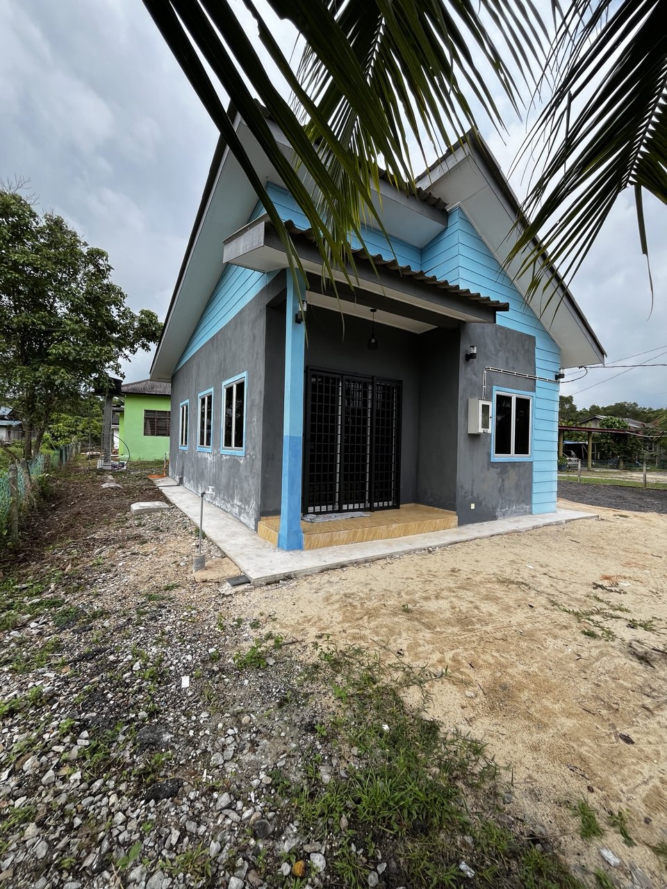 A modern house is depicted with a combination of light blue and gray siding. Two large windows frame the entrance, while a covered porch awaits. Surrounding greenery and gravel pathways lead up to the home, providing a welcoming approach.
