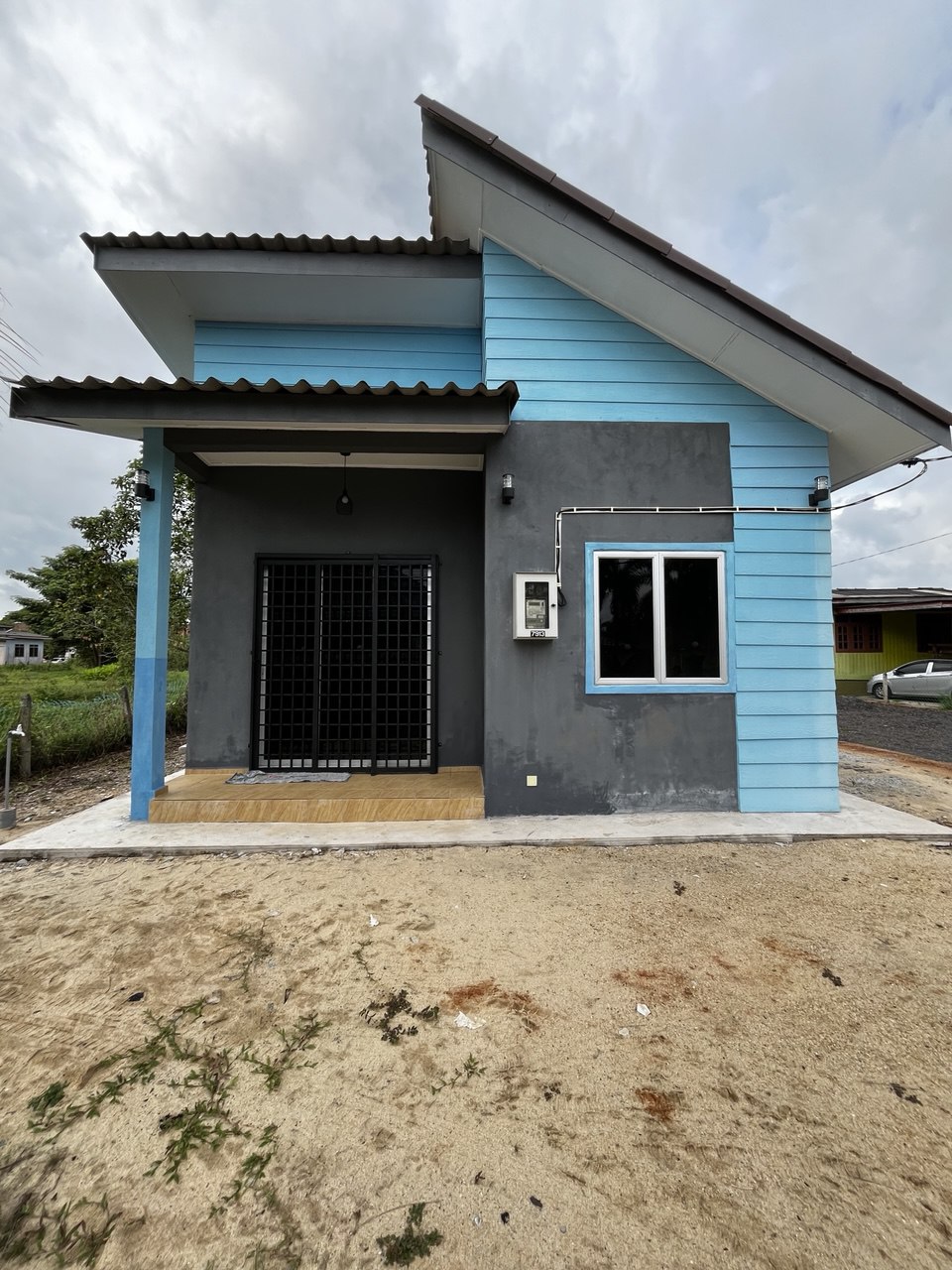 A modern single-story house features a combination of blue and gray siding. A covered porch area with wooden flooring is highlighted by a front door surrounded by a black security gate. Nearby, a gravel path leads to the street, framed by patches of grass.
