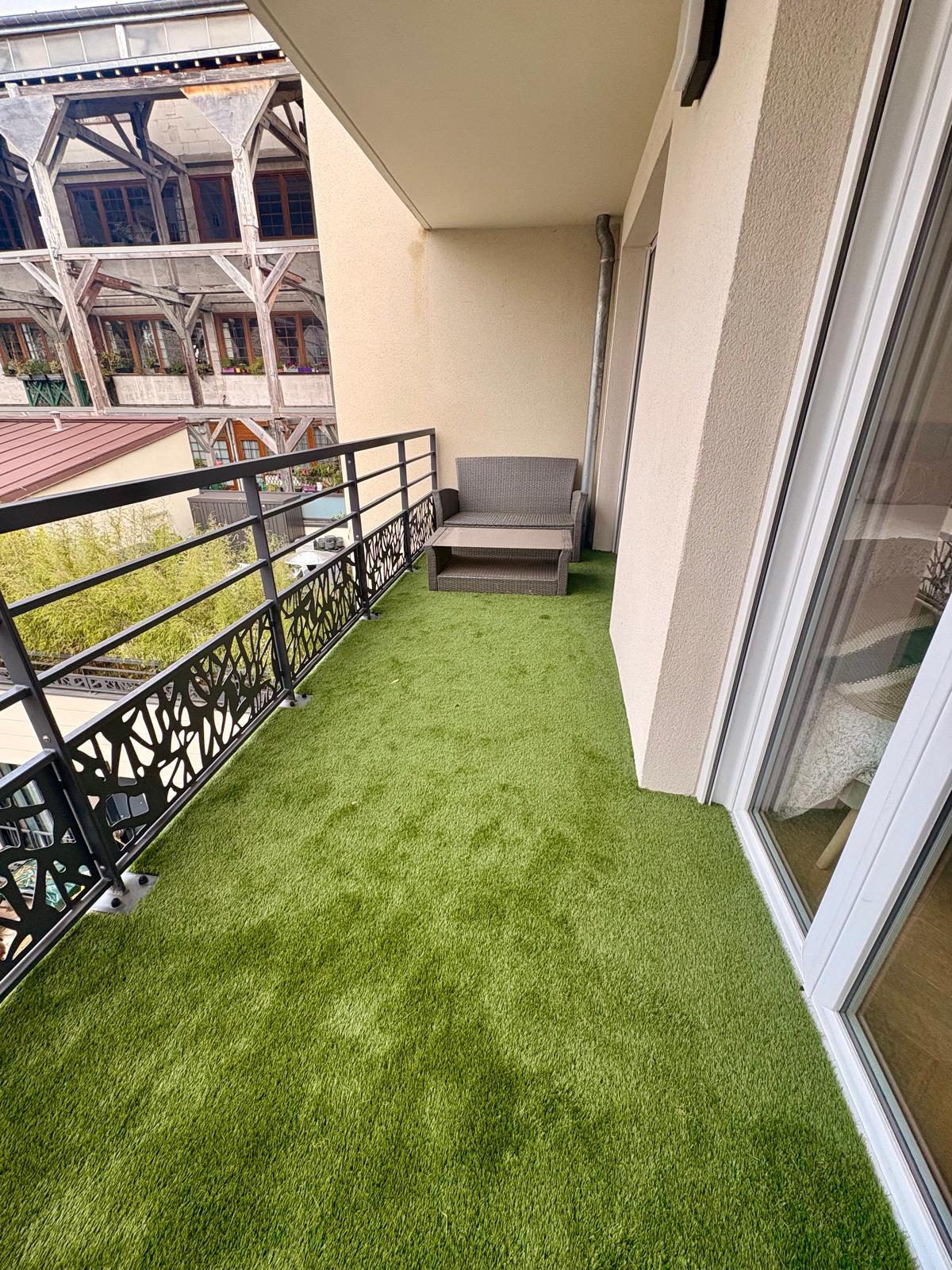 A balcony is shown, featuring a patch of green artificial turf and a single armchair. The railing, designed with a decorative pattern, offers a view of surrounding architectural details. The area appears well-lit and inviting, making it suitable for relaxation.