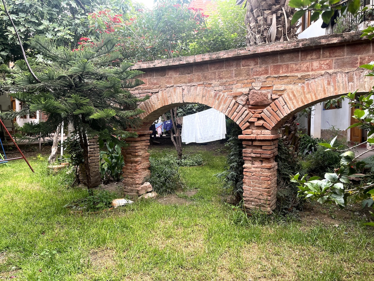 An archway made of brick is seen in a lush garden space, surrounded by greenery. The structure features two robust columns supporting a horizontal beam, creating a sense of openness. In the background, a cloth hangs on a line, subtly contrasting the natural elements.