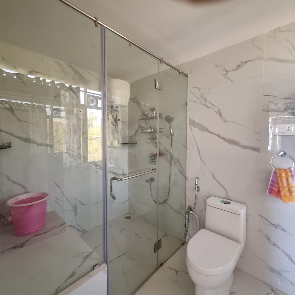 A modern bathroom is highlighted by a glass shower enclosure featuring marble walls and a handheld showerhead. A toilet is positioned to one side, and a pink bucket is partially visible in the corner. Natural light streams in through a window, enhancing the space.