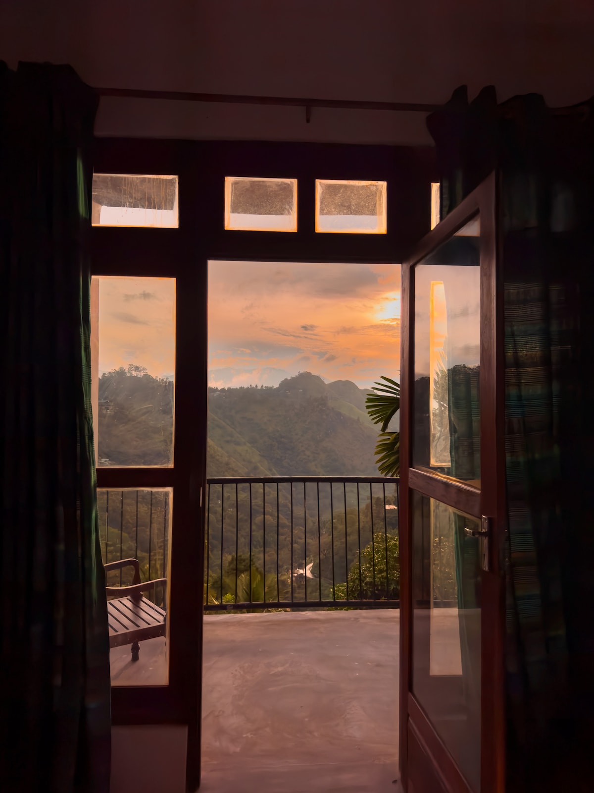 A doorway frames a scenic view of mountains and valleys at sunset. Large glass panels allow natural light to fill the space, while a simple bench is visible on the balcony. Dark curtains gently drape on either side of the entrance.