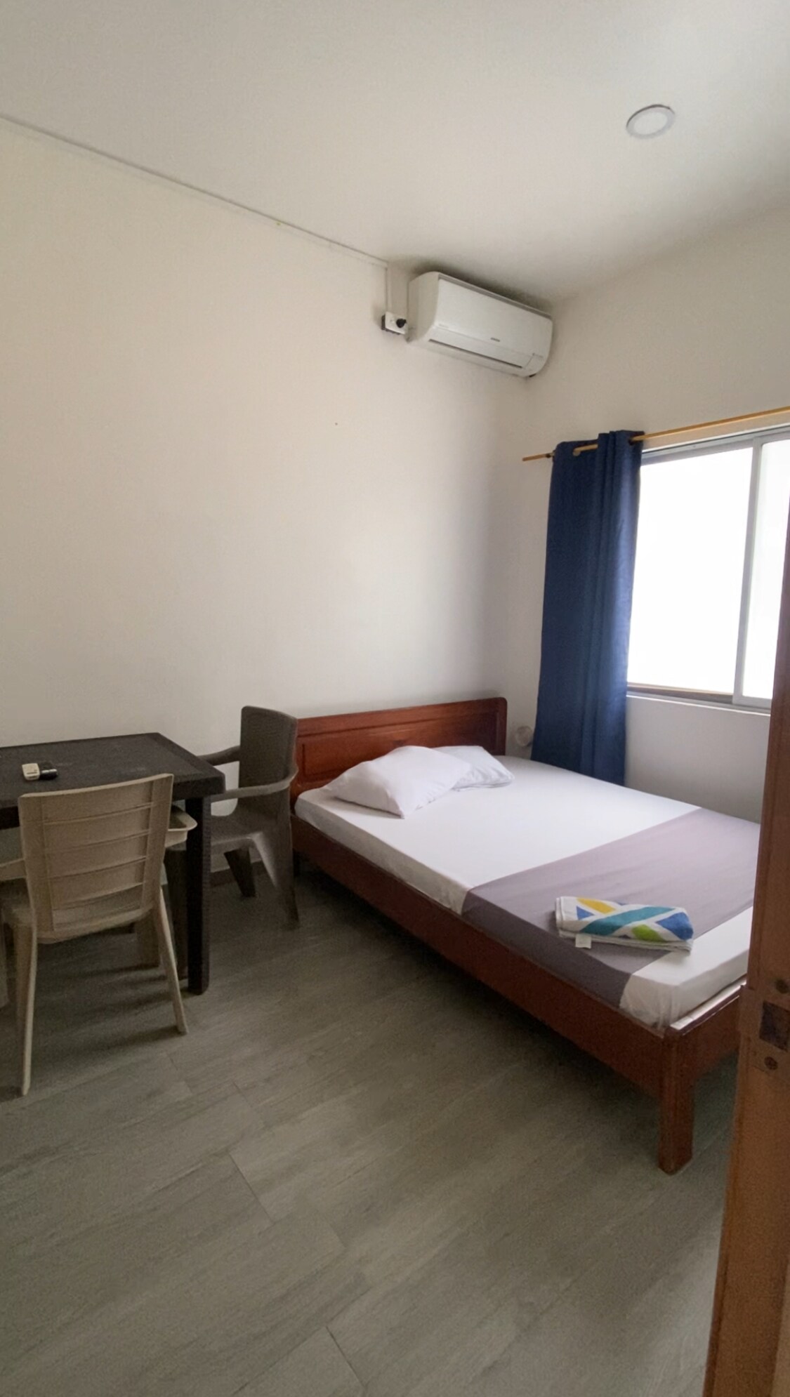 A minimalistic room features a single bed with a white pillow and a folded towel. A small desk and chair are present near a window with dark blue curtains, allowing natural light to filter in. An air conditioning unit is mounted high on the wall.