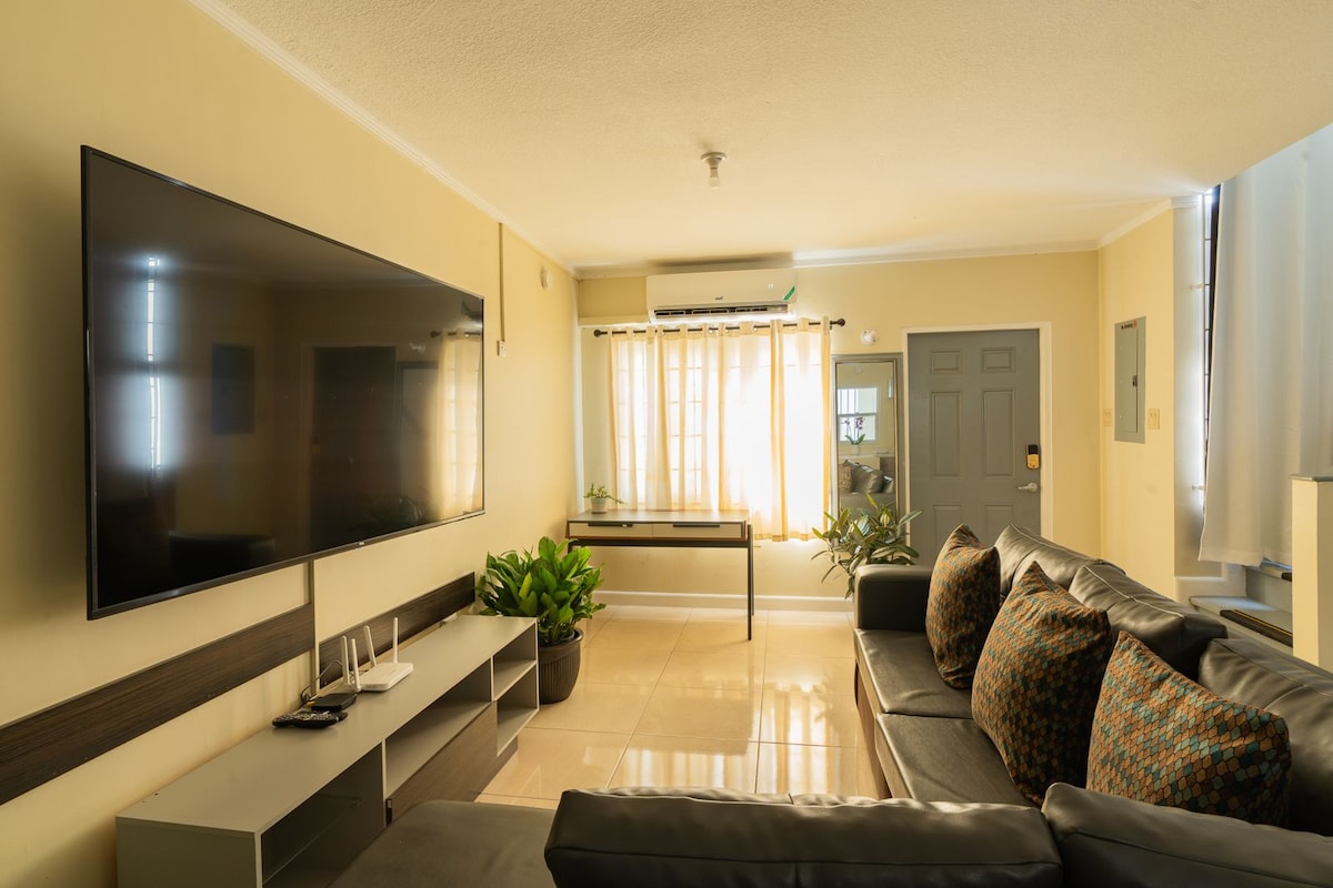 A spacious living area features a large flat-screen television mounted on the wall. A leather sofa with decorative throw pillows is arranged around a coffee table. Natural light floods the room through sheer curtains, with potted plants adding a touch of greenery.