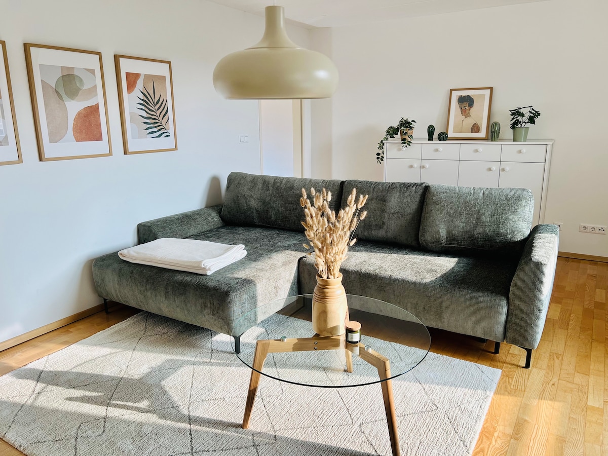 A modern living room is presented with a comfortable gray sofa and a glass coffee table. Framed art pieces adorn the wall above the couch. A decorative vase containing dried flowers adds a subtle touch, while a white throw blanket is neatly placed on the sofa.