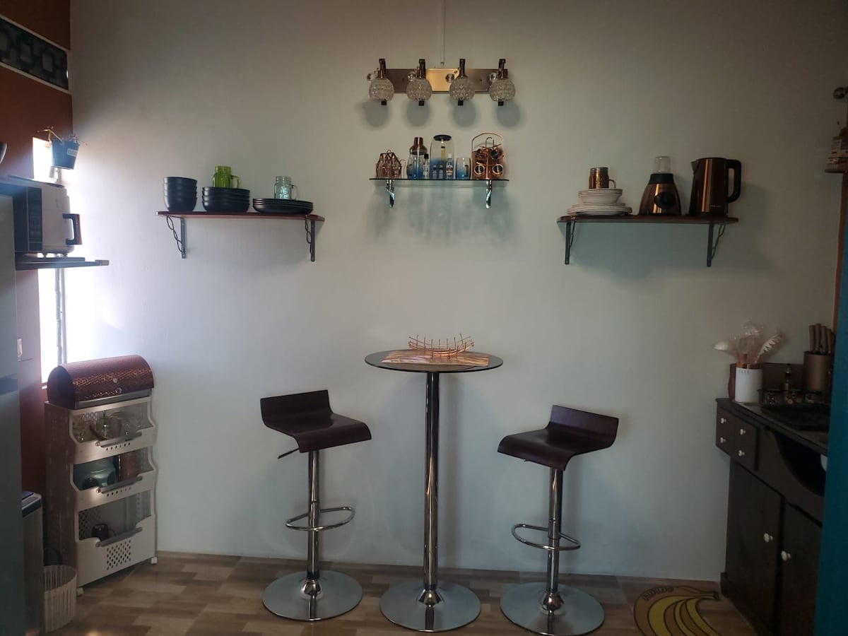 A kitchen area features modern bar stools beside a sleek circular table. Open shelves display various dishes, glassware, and kitchen appliances. A small countertop houses additional kitchen items, emphasizing functionality and style in a compact space.