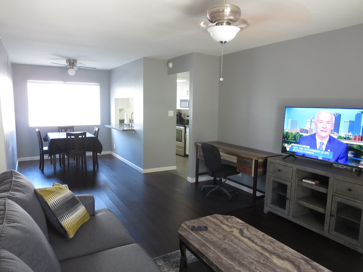 A spacious living area is highlighted by a large window allowing natural light to fill the room. A comfortable gray sofa faces a television on a wooden stand, while a dining table and chairs are visible in the background, along with an open kitchen.