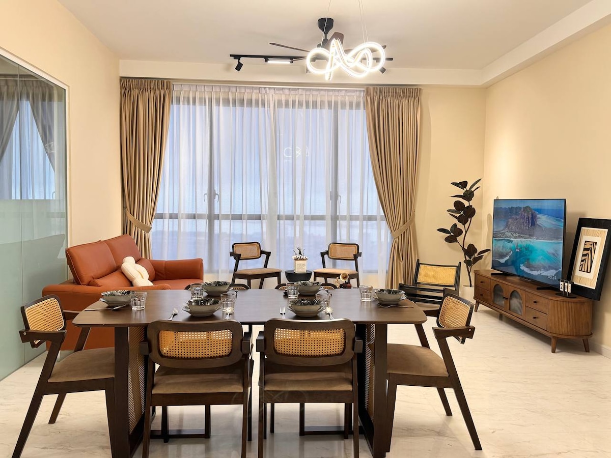 The dining area features a dark wooden table set for six, surrounded by rattan-style chairs. A comfortable orange sofa is positioned near large windows, offering natural light. A flat-screen TV rests on a mid-century modern console, complemented by indoor plants adding a touch of greenery.