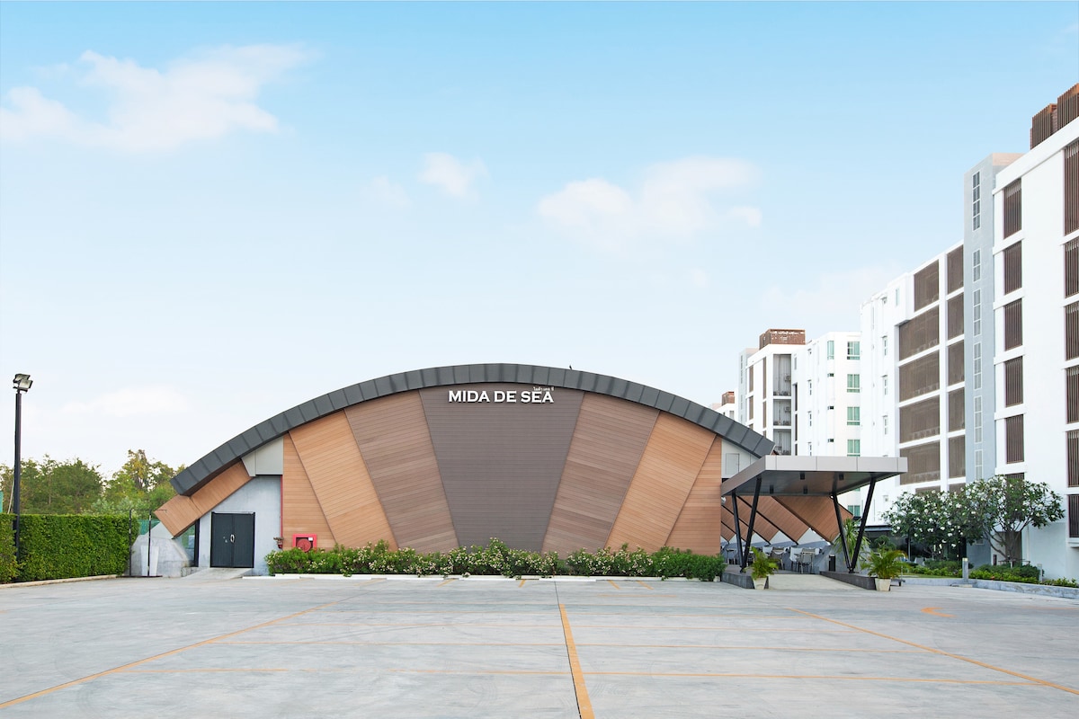 The exterior of Mida De Sea features a modern, curved architectural design with warm wooden accents. The entrance is framed by landscaped greenery and a paved parking area, offering convenient access. The surrounding buildings provide a contemporary backdrop against a clear blue sky.