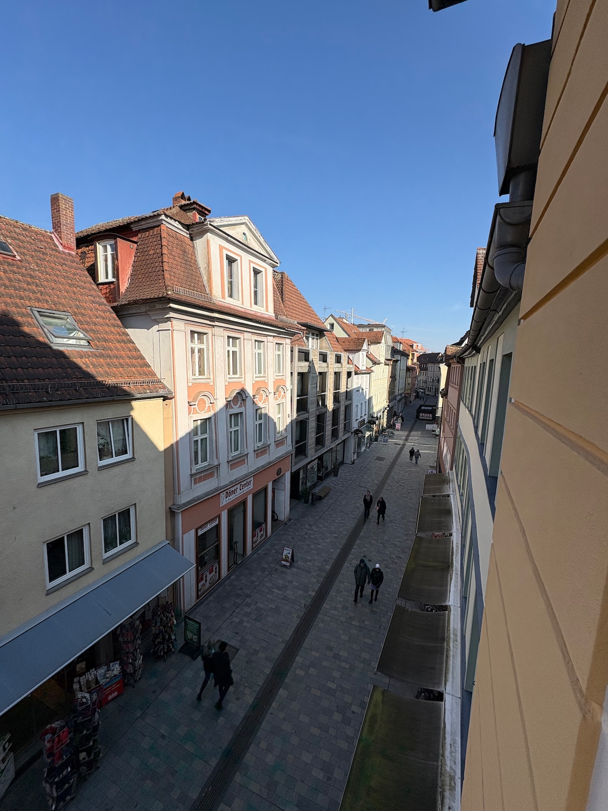 A view from an upper floor showcases a lively street in Ansbach. Modern and historic buildings line the cobblestone path, where pedestrians can be seen strolling. Bright blue skies provide a clear backdrop, enhancing the vibrant atmosphere of the city center.