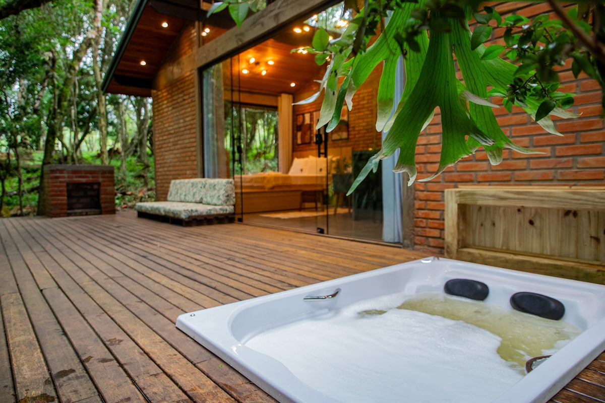 A private outdoor deck features a heated hydromassage bathtub, surrounded by lush greenery. The deck is made of wooden planks, enhancing the rustic charm, while a cozy seating area is visible in the background through large glass doors.