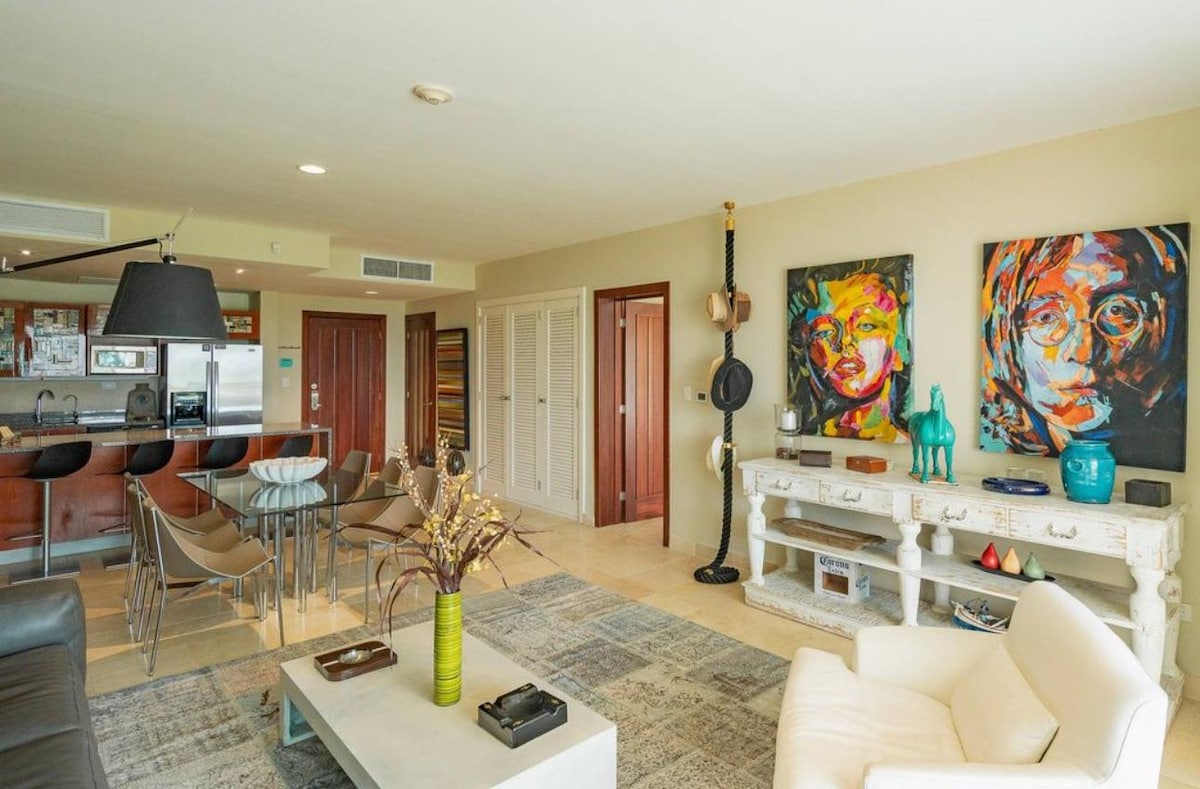 A modern living space is displayed, featuring a light-colored sofa and a white armchair. Decorative wall art adds color to the light walls. A dining area with sleek chairs is paired with a kitchen in the background, equipped with essential appliances.