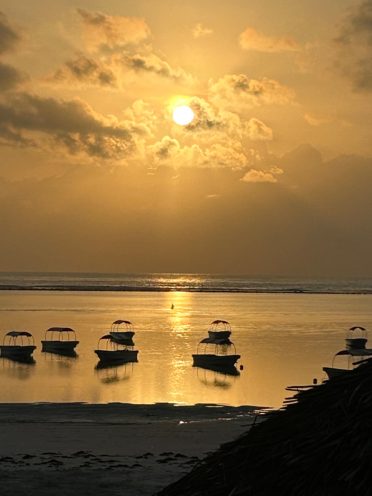 The image captures a serene coastal sunset, with the sun lowering on the horizon, casting a golden hue over the calm water. Several boats are anchored in the foreground, their silhouettes reflecting softly on the shimmering surface, while clouds add texture to the sky.