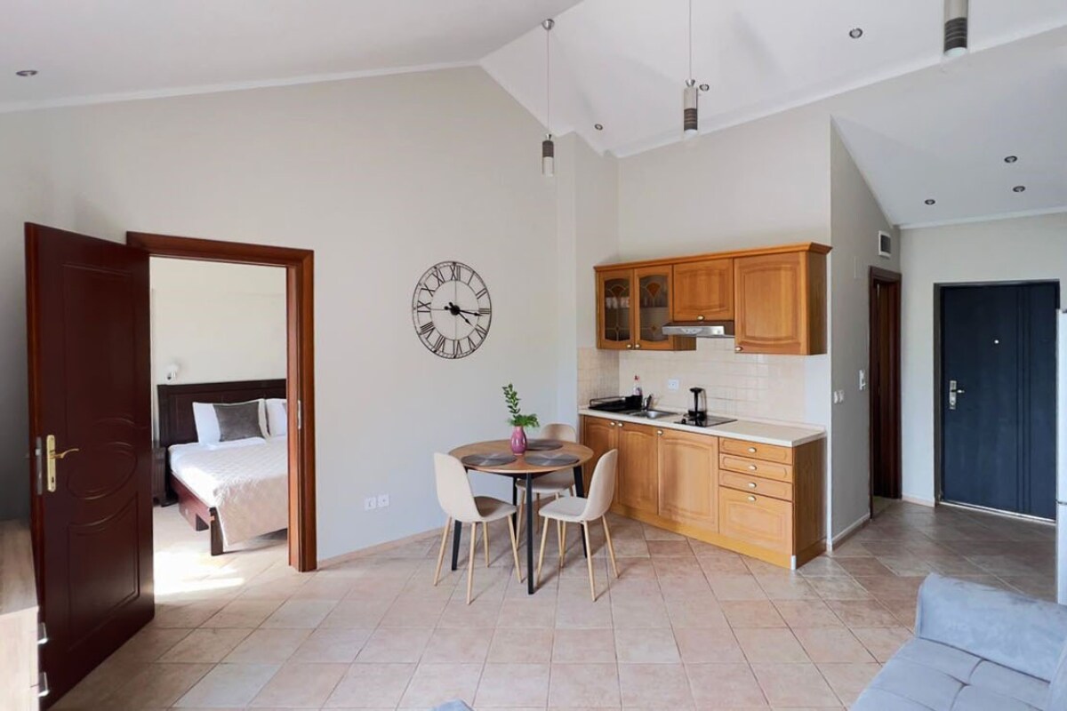 The living space features a compact kitchen with wooden cabinetry and modern appliances. A round dining table with four chairs is positioned nearby. Natural light enters through the doorway, illuminating the adjacent bedroom with a stylish clock on the wall.