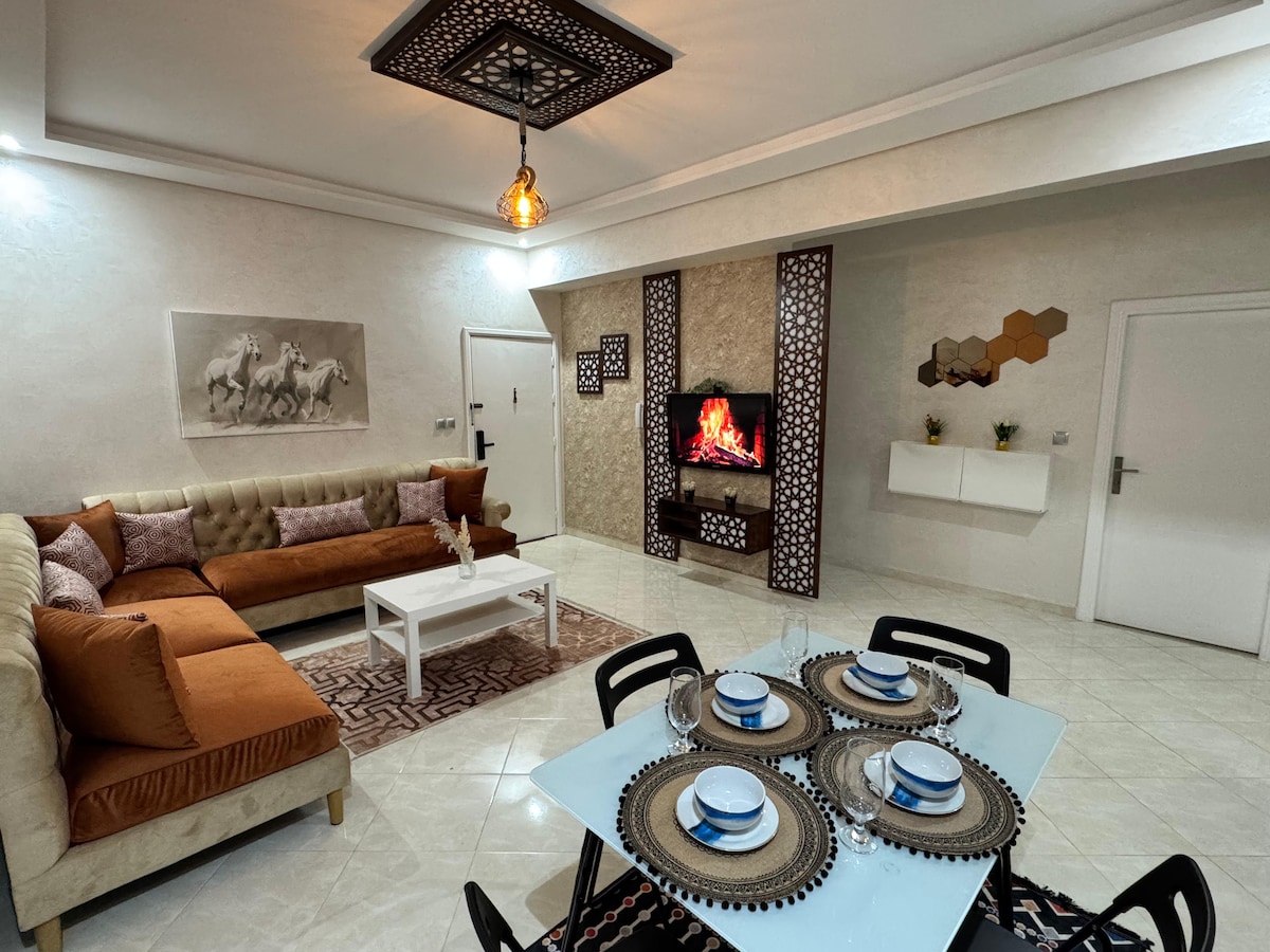 A bright living area is showcased, featuring a large sectional sofa in shades of gold and brown. A modern dining table is set for six, complete with placemats and dishes. A decorative wall unit displays intricate patterns, while a faux fireplace adds warmth to the room.
