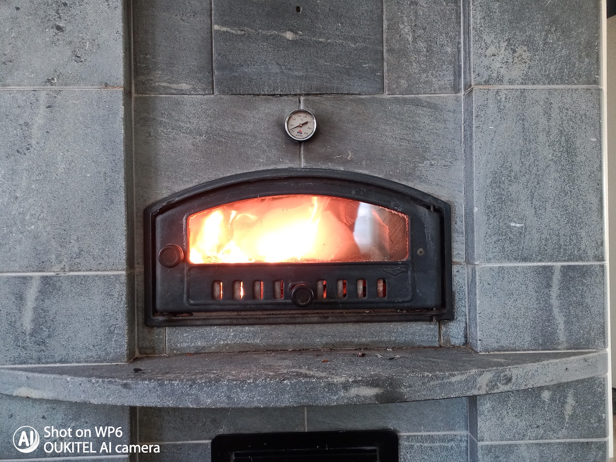 A traditional wood-fired oven is shown, featuring a large arched opening with flames visible inside. The oven's exterior is made of stone, with a circular thermometer mounted above the door, indicating the heat level.