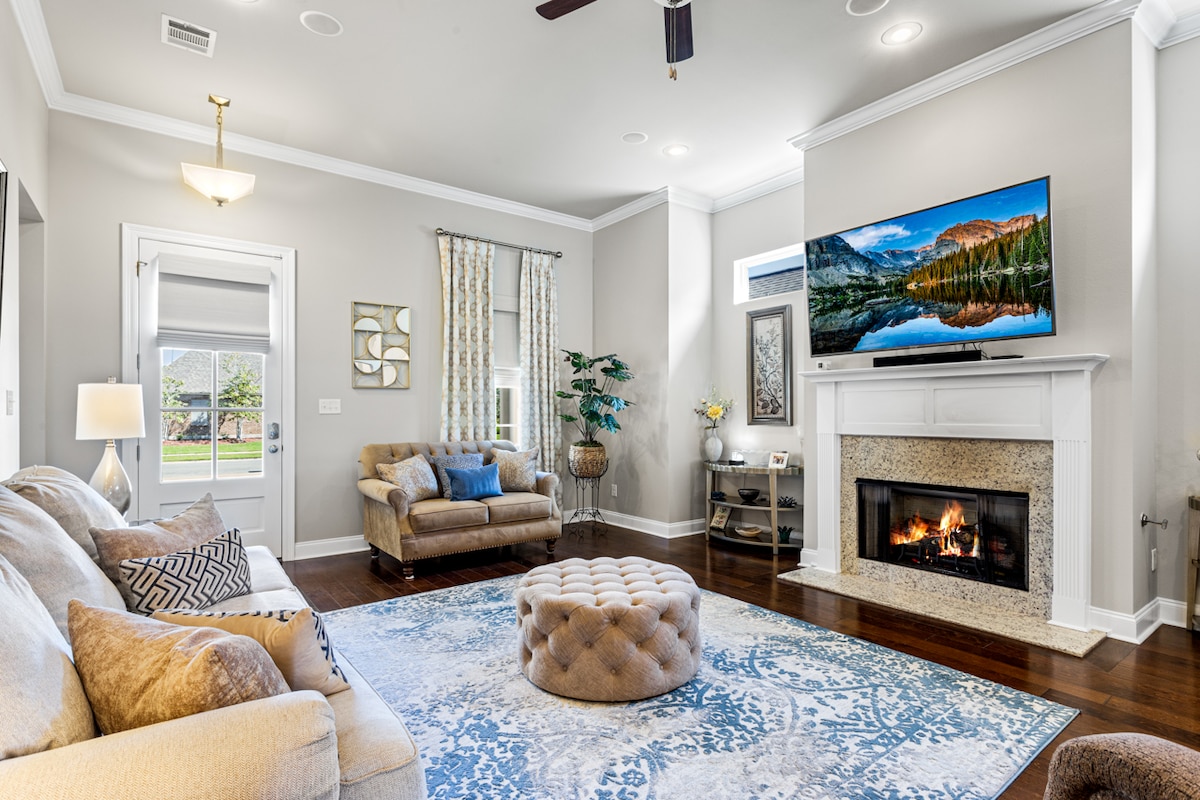 A spacious living area features a large sectional sofa and an accent chair, oriented towards a built-in fireplace with a TV mounted above. A decorative ottoman sits at the center of a soft area rug. Natural light streams through the window, illuminating the room.