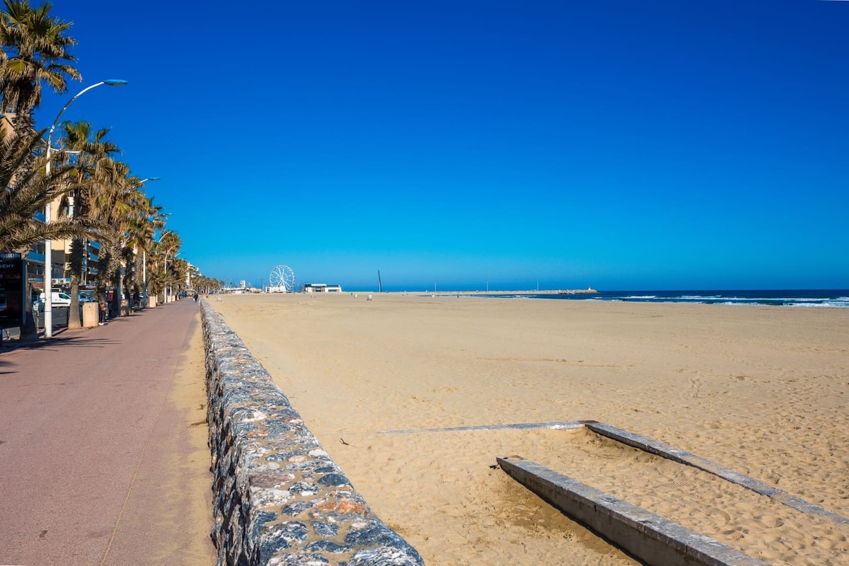 A sandy beach stretches along the shoreline, bordered by a wide stone wall. Palm trees line a smooth pathway next to the beach, while a clear blue sky and ocean create a serene backdrop. A beachfront area is visible, showcasing expansive open space.