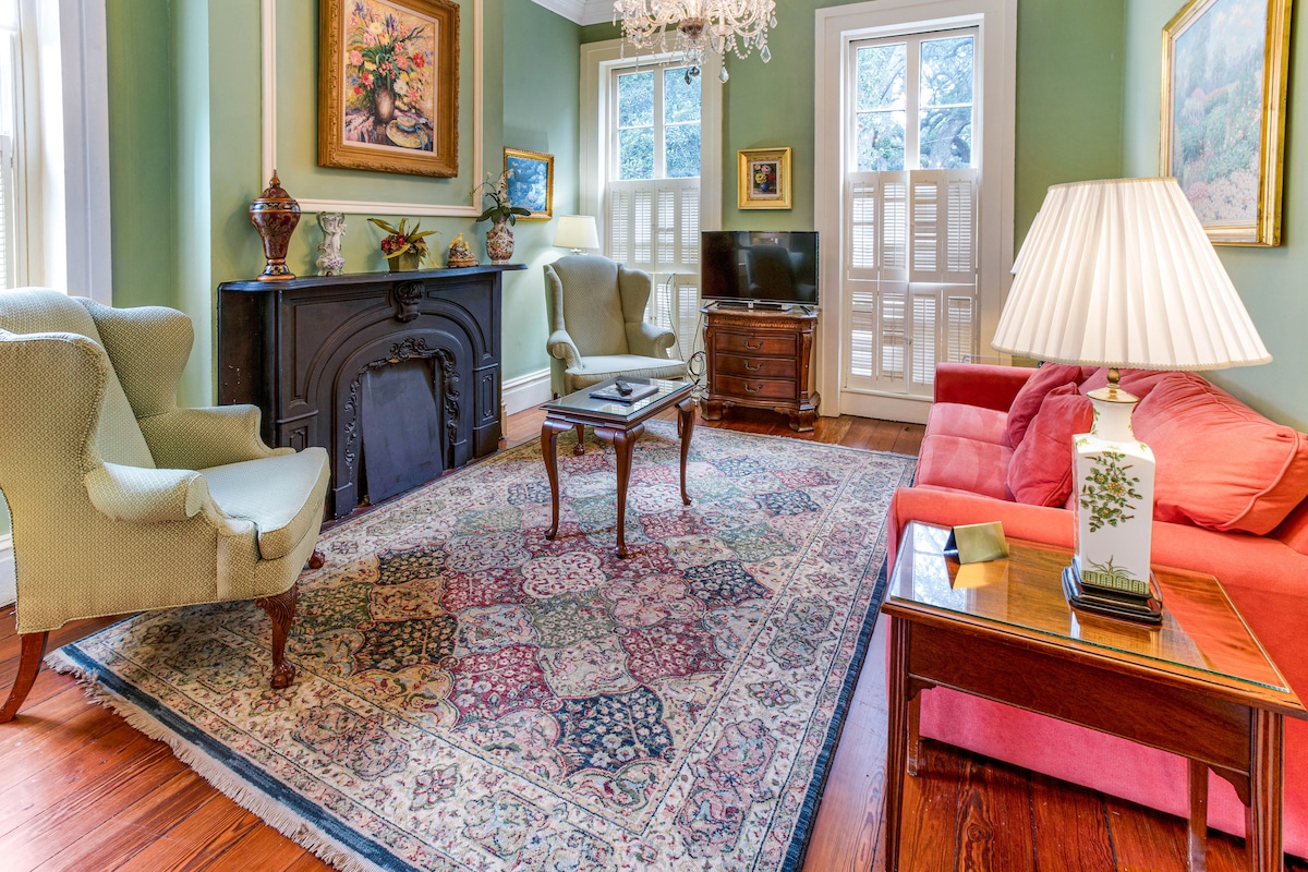 The living room features a warm color palette with a vibrant area rug set on polished wooden flooring. Comfortable seating options include two armchairs and a sofa. Natural light filters in through windows adorned with shutters, while a television and decorative elements complete the space.