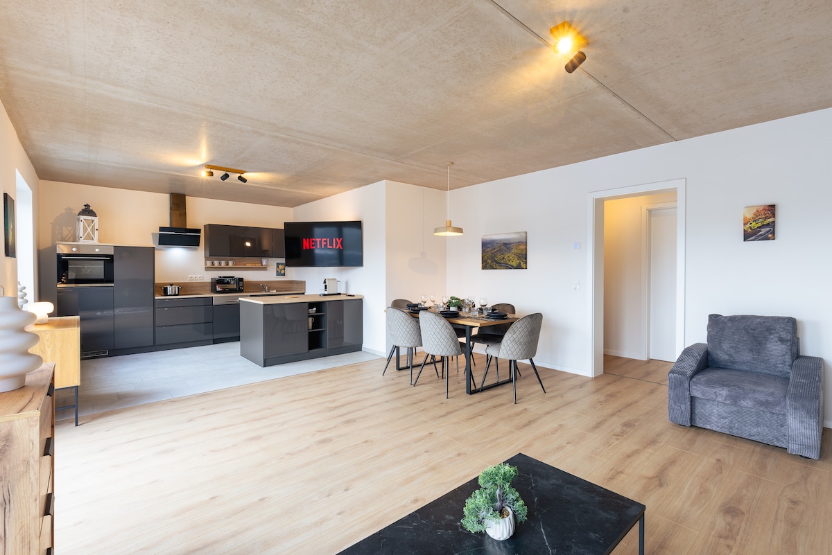 An open layout showcases a modern kitchen with dark cabinetry and a breakfast bar. A dining table with chairs is positioned nearby, with a cozy gray armchair situated in the corner. Natural light fills the space, highlighting the warm wooden flooring.