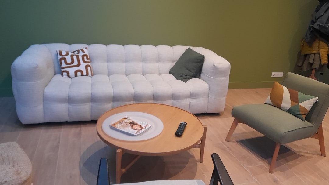 A light-toned, tufted sofa is placed against a green wall, complemented by a decorative throw pillow. A round wooden coffee table is positioned in front, featuring a magazine and a remote control. A small green armchair with a patterned cushion completes the seating area.