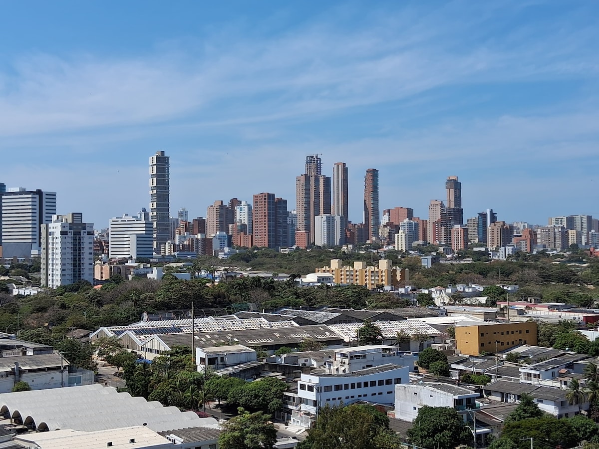 A panoramic view of the city skyline is presented, featuring several high-rise buildings and a mix of green spaces. The foreground shows low-rise structures and rooftops, offering a contrast to the taller urban backdrop under a clear blue sky.