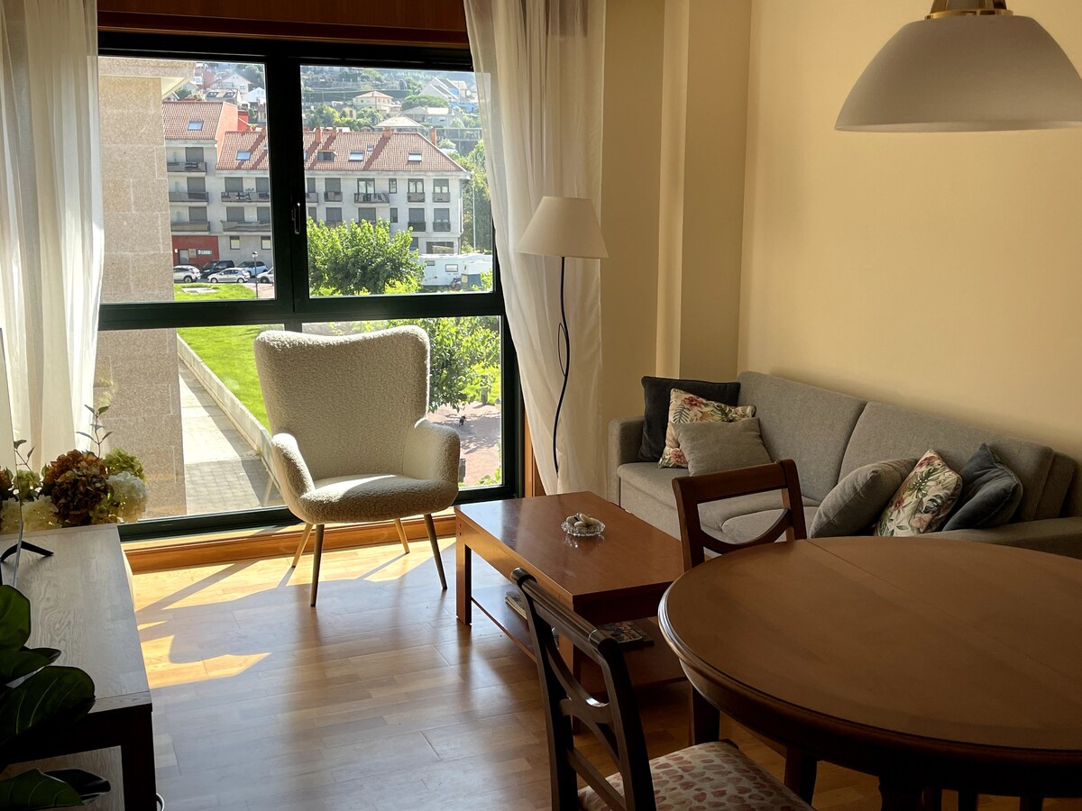 A well-lit living area features a comfortable couch and an inviting armchair positioned near a large window. The wooden dining table is set for conversation. Soft curtains frame the window, allowing natural light to enhance the space while offering views of the surrounding greenery.