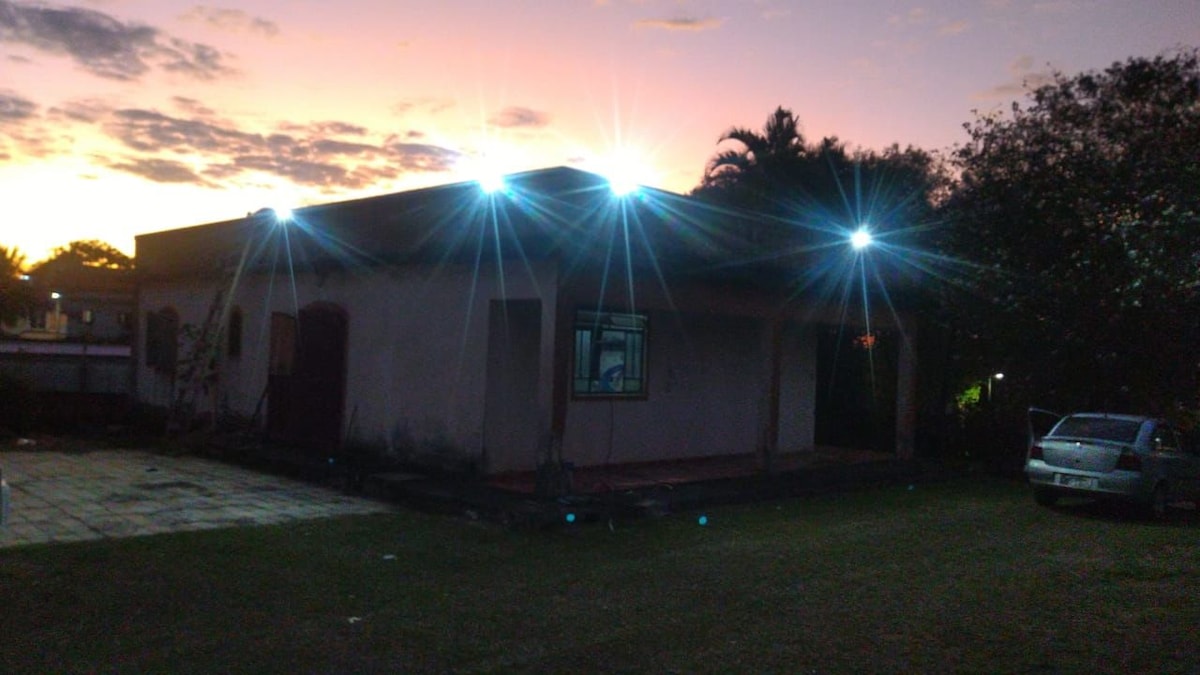 The exterior of the house is shown during twilight, illuminated by bright lights atop the structure. A spacious grassy area surrounds the building, with a vehicle parked nearby. The sky features soft hues of orange and purple, adding a serene backdrop.