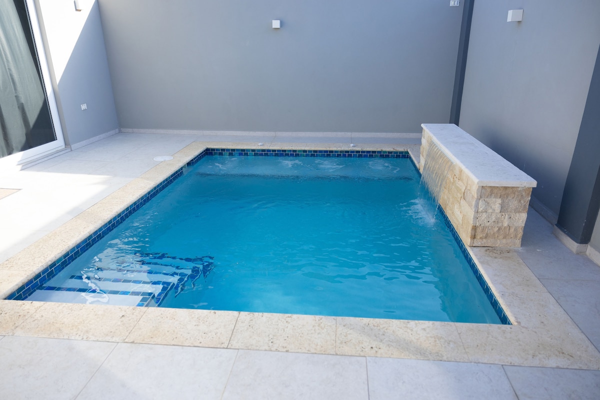 A private pool is situated within a stone-paved area, featuring a clear blue surface and a textured stone wall at one end. The surrounding walls are painted in a soft gray, providing a serene setting.