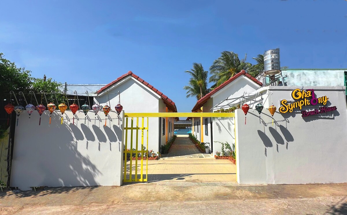 The entrance is framed by a colorful gate leading to the pathway, flanked by two charming white structures with terracotta roofs. Lush greenery and decorative lanterns add vibrant accents, while the ocean can be glimpsed in the background, enhancing the welcoming ambiance of the setting.