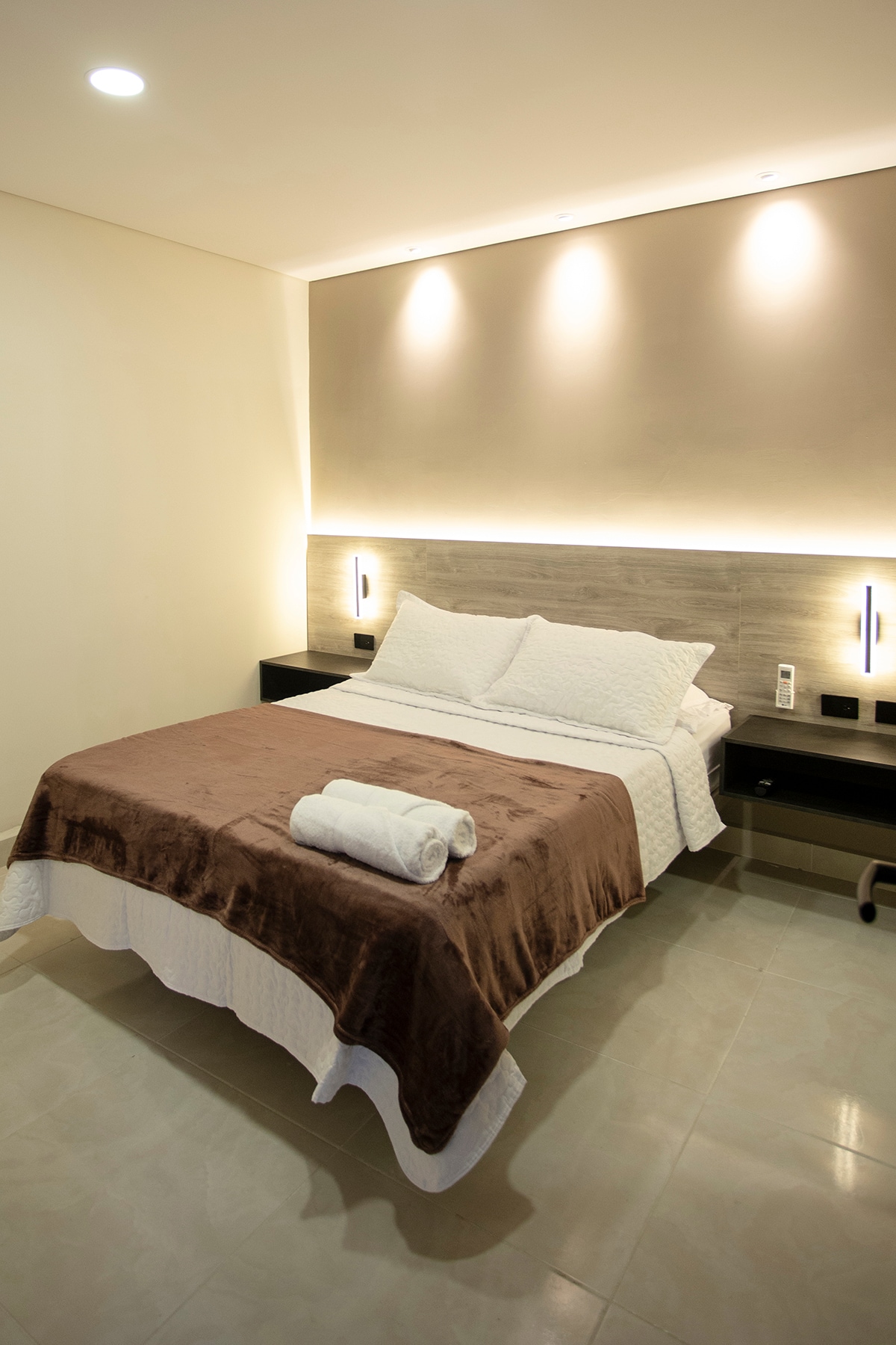 A spacious bedroom is furnished with a large bed covered with a smooth, light-colored quilt and a brown throw. Soft lighting highlights the headboard and bedside tables. Two rolled towels are neatly placed on the bed's edge, complementing the modern, minimalist design.