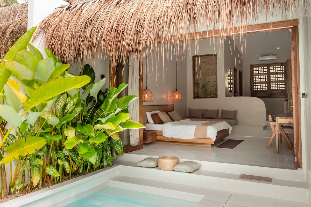 A serene bedroom space features a spacious bed adorned with neutral-toned linens. Natural light pours in through an opening, illuminating the area. Lush green plants are positioned near the entry, complementing the thatched roof and enhancing the peaceful ambiance.